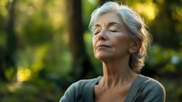 Mature woman with closed eyes enjoying peaceful moment in nature. Golden sunlight illuminates her face while gentle breeze moves through silver hair. Serene outdoor meditation.