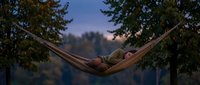 Relaxed man resting in hammock suspended between trees at dusk, surrounded by forest foliage and peaceful natural landscape for wellness and outdoor lifestyle content.
