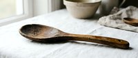 Rustic wooden spoon with weathered patina resting on white linen cloth in farmhouse kitchen setting with ceramic bowls creating cozy culinary atmosphere.