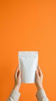 Hands holding blank white pouch against vibrant orange background for product packaging mockup and branding presentations.
