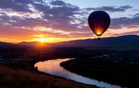 Hot air balloon floating over scenic valley landscape during golden hour sunrise with river and mountains creating dramatic silhouette against colorful sky.
