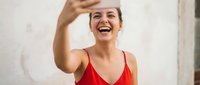 Happy young Caucasian woman in red top taking selfie with smartphone, laughing joyfully while capturing self-portrait for social media sharing.