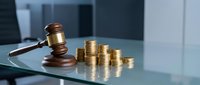 Wooden judge gavel next to stacks of golden coins on glass table in modern office setting representing legal fees, court costs, and financial justice.