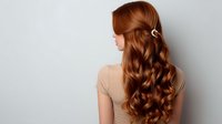 Young woman with long wavy auburn hair styled with elegant clip shown from behind against light background, demonstrating professional hairstyling and healthy hair texture.