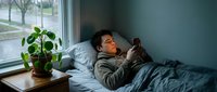Young Caucasian male teen lying in bed using smartphone, face lit by screen glow, cozy bedroom with window and houseplant.