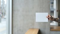 Tattooed male hand holding blank white card in modern urban loft interior with concrete walls and natural window light for mockup and branding purposes.