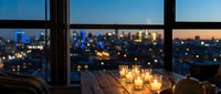 Cozy evening scene with glowing candles on wooden table by large windows overlooking illuminated city skyline at dusk creating warm romantic atmosphere.