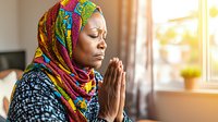 Young African American Muslim woman in colorful hijab praying peacefully by window with sunlight, eyes closed and hands clasped in spiritual meditation moment.