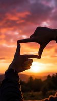 Hands forming frame gesture against dramatic sunset sky with orange and purple clouds over mountain landscape silhouette for creative vision concepts.