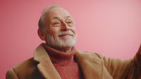 Senior Caucasian man with white beard wearing turtleneck sweater and beige coat smiling warmly against pink background, expressing joy and contentment in lifestyle portrait.
