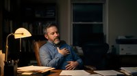 Middle-aged Caucasian businessman working late at office desk with warm lamp lighting, thoughtful expression while reviewing documents in corporate environment.