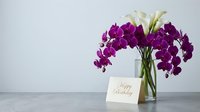 Purple orchids and white calla lilies in glass vase with happy birthday card on marble surface against white background for greeting celebrations.