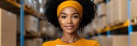 Young African American woman warehouse worker in orange uniform and headband smiling confidently in modern logistics facility with shelving systems.
