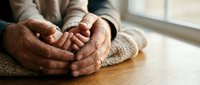 Elderly hands clasped together in prayer or meditation pose on wooden table by window, conveying peace, spirituality, and contemplation in warm natural light.