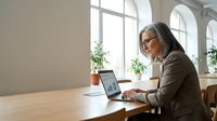 Senior businesswoman with gray hair working on laptop in modern office with large windows and natural light for professional corporate content.