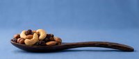 Wooden spoon filled with mixed nuts including cashews, almonds, walnuts and raisins on blue background for healthy snacking and cooking concepts.