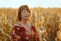 Middle-aged Caucasian woman with curly red hair wearing floral dress standing in golden cornfield at sunset, eyes closed peacefully in natural outdoor setting.