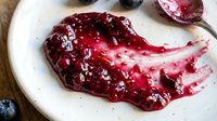 Homemade berry jam with glossy texture spread on white plate, featuring deep red and burgundy tones with fresh fruit pieces, ideal for food styling and culinary content.