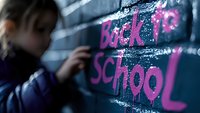 Back to school graffiti text in pink spray paint on dark wall with blurred student silhouette in foreground creating atmospheric education concept.