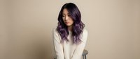 Young Asian woman with purple hair wearing white sweater looking down pensively against neutral beige background for lifestyle and fashion concepts.