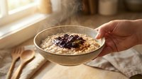 Hand holding bowl of steaming oatmeal porridge topped with berries and cinnamon for breakfast, healthy eating and morning meal preparation.