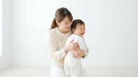 Young Asian mother in cream sweater tenderly holding crying baby in white outfit against minimalist background for parenting and childcare content.