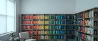 Modern home library with colorful books arranged by rainbow spectrum on wooden shelves, minimalist reading corner with chair and natural window light.