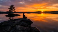 Woman in silhouette meditating on rocky shore at golden sunset, peaceful lakeside scene with calm water reflections and solitary tree, mindfulness and tranquility concept for wellness.