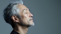 Serene mature Asian man with gray hair and beard in profile view against gray background, eyes closed in peaceful meditation or mindfulness moment.