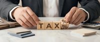 Male professional in dark business suit arranging wooden blocks spelling tax with percentage symbol on desk, representing tax planning and financial management services.