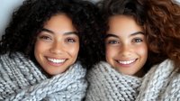 Two happy young women with curly hair wrapped in cozy gray knit blankets smiling warmly at camera. Perfect for friendship, comfort, wellness content.