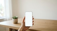 Hand holding smartphone with blank white screen on wooden desk with potted plant and window light in background.