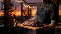 Professional chef presenting grilled steak on wooden board during golden hour barbecue session with warm string lights creating cozy outdoor dining atmosphere.