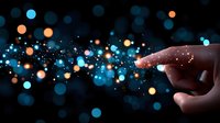 Magical glowing particles flow from pointing finger silhouette against dark background with blue and orange bokeh lights creating ethereal atmosphere.
