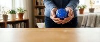 Person holding blue smiley face ball in cupped hands at wooden table in cozy home interior with plants and bookshelves for positive mindset and wellness.