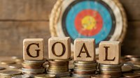 Wooden blocks spelling GOAL stacked on coin piles with archery target in background, symbolizing financial planning and success.