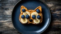 Creative breakfast toast shaped as cat face with peanut butter, banana slices and fresh blueberries served on dark plate against rustic wooden background.