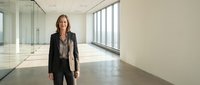 Mature woman with long brown hair, Caucasian ethnicity, wearing dark business blazer and pants, standing in modern empty office space with floor-to-ceiling windows and polished concrete floors, suitable for corporate, leadership, and business concepts.