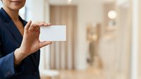 Mature woman in navy blazer holding blank white business card in modern office interior, suitable for corporate branding and professional networking materials.