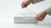 Hands unwrapping clear plastic film from white cardboard box on neutral background, packaging and delivery concept.