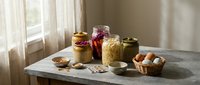 Homemade preserves and pickled vegetables in glass jars on rustic wooden table near window with natural light for pantry storage and cooking.