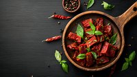 Sun-dried tomatoes with fresh basil leaves in wooden bowl on dark background, seasoned with black pepper and spices, top view food photography.