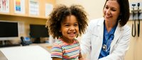 Happy African American girl with curly hair smiling during pediatric checkup with female doctor in medical office. Healthcare, child wellness, medical examination.