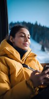 Young woman in yellow winter jacket relaxing by window during mountain trip, snowy landscape visible outside, enjoying peaceful moment.