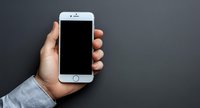 Male hand holding modern smartphone with blank black screen against dark gray background, wearing light blue business shirt sleeve. Mockup for app or web design.