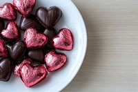Heart shaped chocolate candies in dark brown and metallic pink foil wrapping on white plate against neutral background, romantic holiday confectionery setting.
