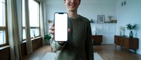 Smiling young Caucasian man in green sweater holding smartphone with blank white screen mockup toward camera in modern living room.