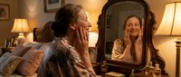 Young woman applying skincare cream while looking in vintage mirror at cozy bedroom vanity with warm lighting for beauty and self-care routine.