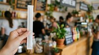 Young adult with medium skin tone holding blank white paper roll in plant shop interior, displaying product packaging or promotional material for retail, gardening, or eco-friendly business concepts.