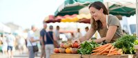 Young woman shopping for fresh organic vegetables at outdoor farmers market with colorful produce display and green umbrellas in background.